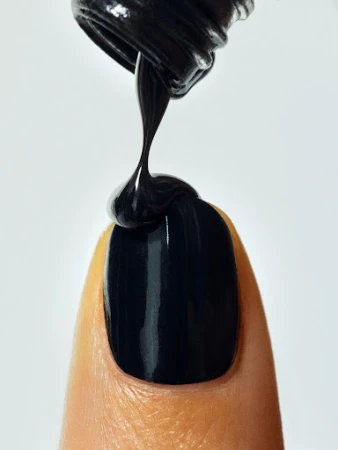 Close-up of black nail polish being applied to a fingernail, set against a clean white background with focus on the polish application.