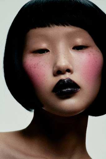 Portrait of an Asian adult female with glossy black lipstick, heavy pink blush, and a bob haircut against a white background.