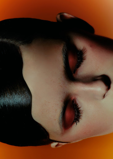 Close-up portrait of a woman with bold red eyeshadow and black hair against a solid orange background, focusing on beauty and makeup artistry.
