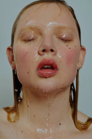Close-up portrait of a woman with wet skin, slicked hair, and closed eyes against a neutral background by Zoe Kovacs
