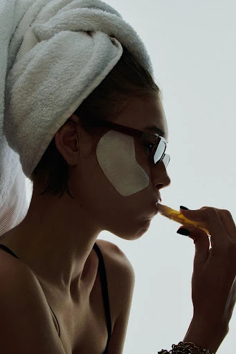 Young woman wearing a towel on her head and eye patches applies beauty product with applicator, indoors against a white background.