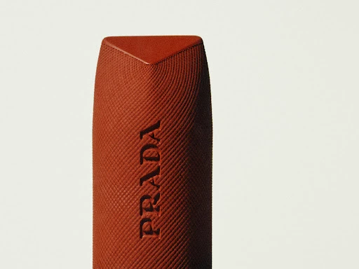 Close-up of Prada lipstick in a warm brown tone with a triangular top and textured body, set against a minimalist off-white background.