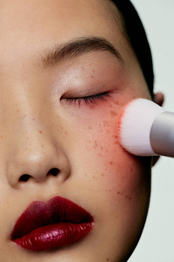Asian adult female having makeup applied with a white makeup brush. She has dark red lipstick and rosy blush.