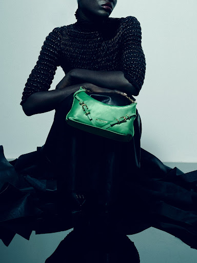 Dark-skinned fashion model poses with a green leather purse in a photo studio, wearing a high-necked top and a draped fabric skirt.