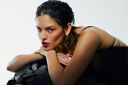 Portrait of a young adult female posing with wet hair, wearing a brown dress and green earrings against a white backdrop in a photo studio.