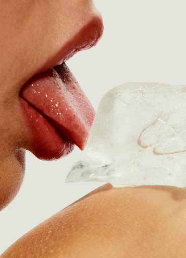 Close-up of a person's face with their tongue touching a block of ice against a white background. Focus on skin and texture.
