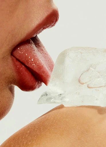 Close-up of a person's face with their tongue touching a block of ice against a white background. Focus on skin and texture.