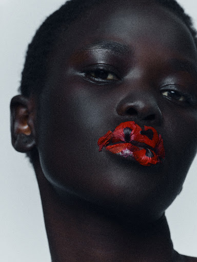 Close-up portrait of a Black woman with short hair wearing red flower petals pressed onto her lips against a neutral background.