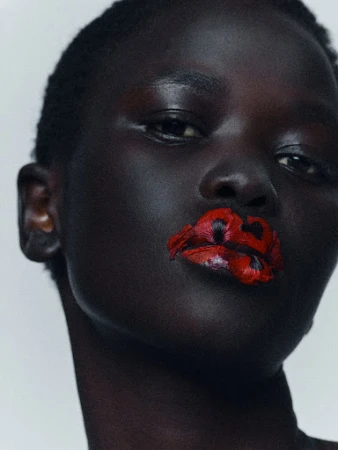 Close-up portrait of a Black woman with short hair wearing red flower petals pressed onto her lips against a neutral background.