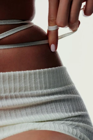 Close-up of a person wearing ribbed white knit shorts with white string ties wrapped around the waist against a white background.