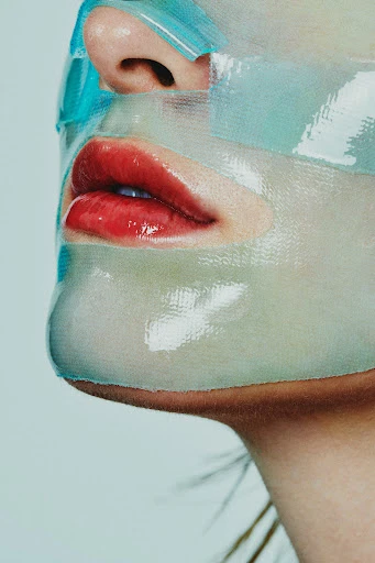 Close-up of a person's face covered in a turquoise sheet mask with glossy red lips, focusing on beauty treatment and skincare.