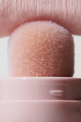 Close-up of a pink makeup brush with soft bristles, positioned on a container of matching light pink color, under bright lighting.
