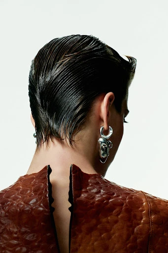 A model from the back wearing a brown textured dress and silver earrings, hair combed back, against a neutral white background. Beauty shot.