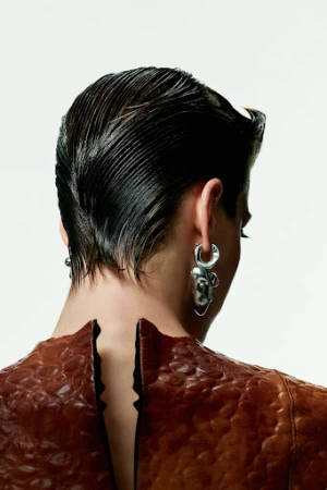 A model from the back wearing a brown textured dress and silver earrings, hair combed back, against a neutral white background. Beauty shot.