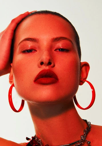 Close-up portrait of a model with a red color grade, featuring red lipstick, red hoop earrings, and a silver chain necklace.