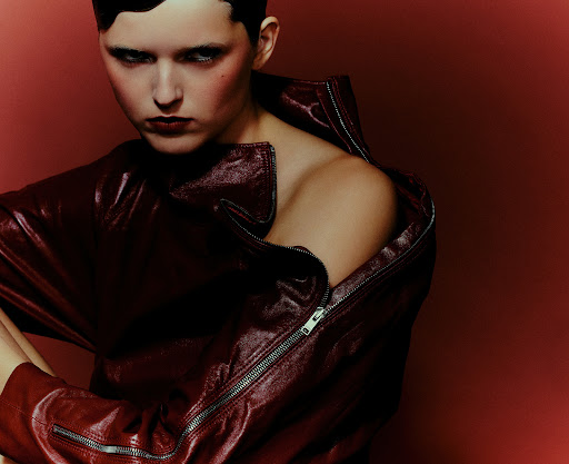 Adult female posing in a red leather jacket with an open zipper. Shot in a studio with a deep red backdrop and dramatic lighting.