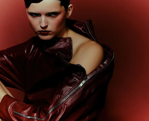 Adult female posing in a red leather jacket with an open zipper. Shot in a studio with a deep red backdrop and dramatic lighting.