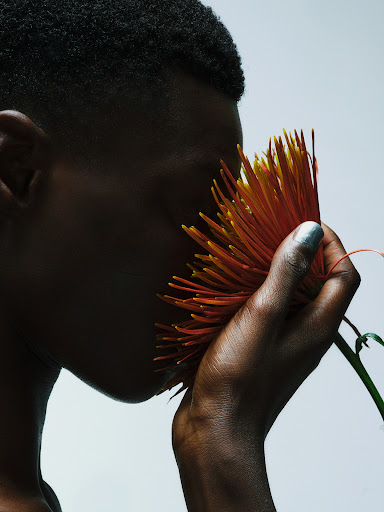 Close-up studio portrait of a dark-skinned adult with short hair, holding a large orange chrysanthemum flower to the face.