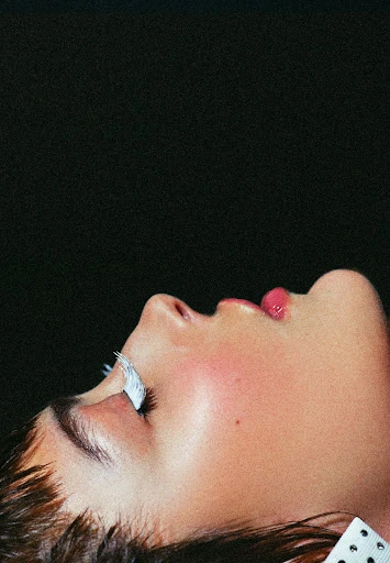 Profile view of an adult female face with pink lips and long lashes against a black background in a beauty or fashion context.