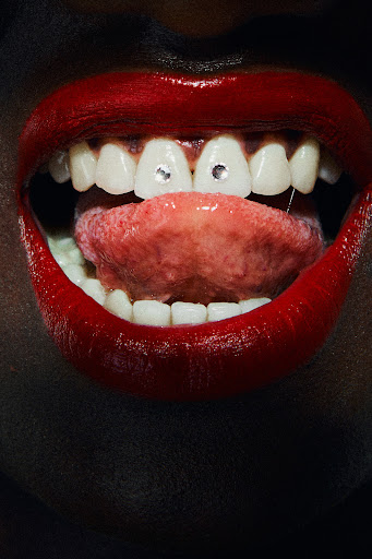 Close-up of a woman's open mouth featuring red lipstick, white teeth with jewel embellishments, and a pink tongue.