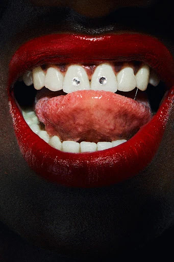 Close-up of a woman's open mouth featuring red lipstick, white teeth with jewel embellishments, and a pink tongue.