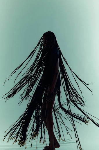 Striking fashion portrait of a Black female model with elaborate braided hair, against a pale teal backdrop, showcasing beauty and style.