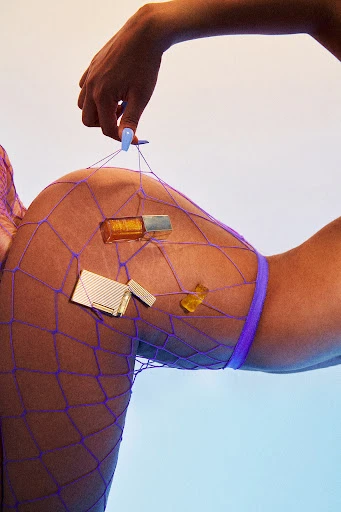 Partial view of a human torso draped in purple fishnet with beauty products attached, against a white and light-blue gradient backdrop.