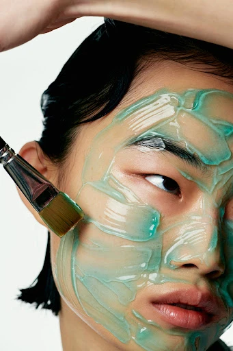 Close-up of a person's face covered with a blue face mask being applied with a brush in a beauty or skincare context.