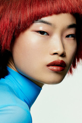 Close-up shot of a person with red hair wearing a blue turtleneck shirt in a studio setting. The focus is on the face and hair.