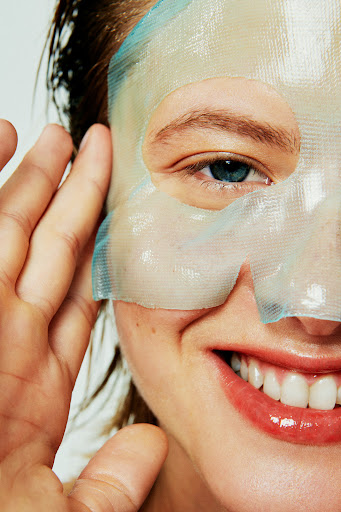 Close-up of a smiling adult woman with a hydrating sheet mask on the left side of her face, her hand near her ear.