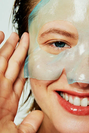 Close-up of a smiling adult woman with a hydrating sheet mask on the left side of her face, her hand near her ear.