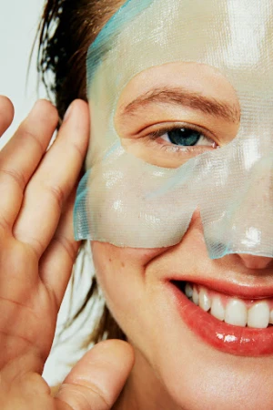 Close-up of a smiling adult woman with a hydrating sheet mask on the left side of her face, her hand near her ear.