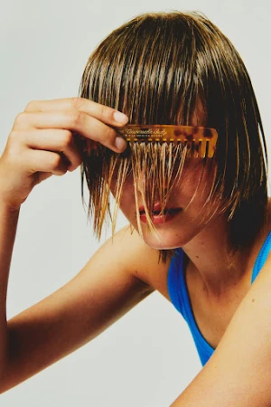 A young adult female with wet short hair poses indoors, holding a tortoiseshell comb to her face against a neutral grey background.
