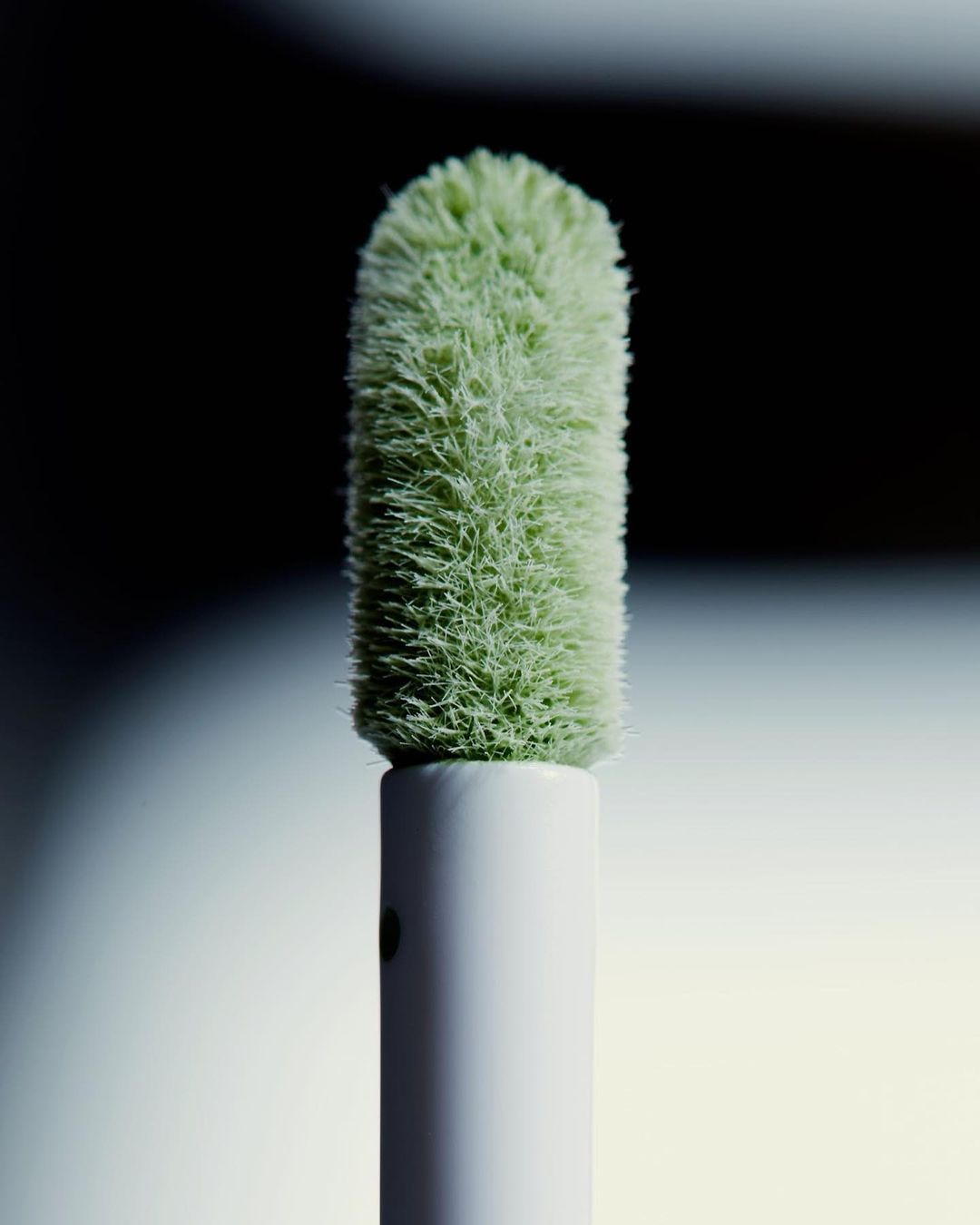 Close-up of a green, flocked cosmetic applicator with a white handle, set against a blurred, dark background for beauty product photography.