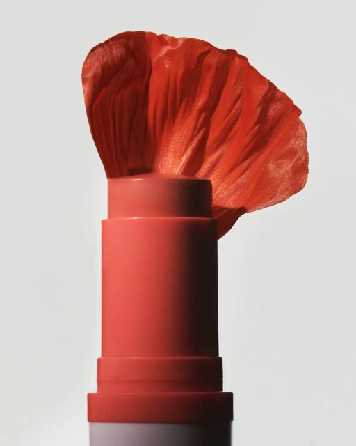 A close-up of a coral-colored lipstick tube topped with an orange flower petal, set against a simple, light gray background.