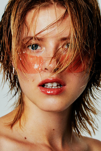 Close-up shot of a woman with wet, short, light-brown hair, clear eye patches, and glossy lips, set against a plain white background.