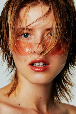 Close-up shot of a woman with wet, short, light-brown hair, clear eye patches, and glossy lips, set against a plain white background.