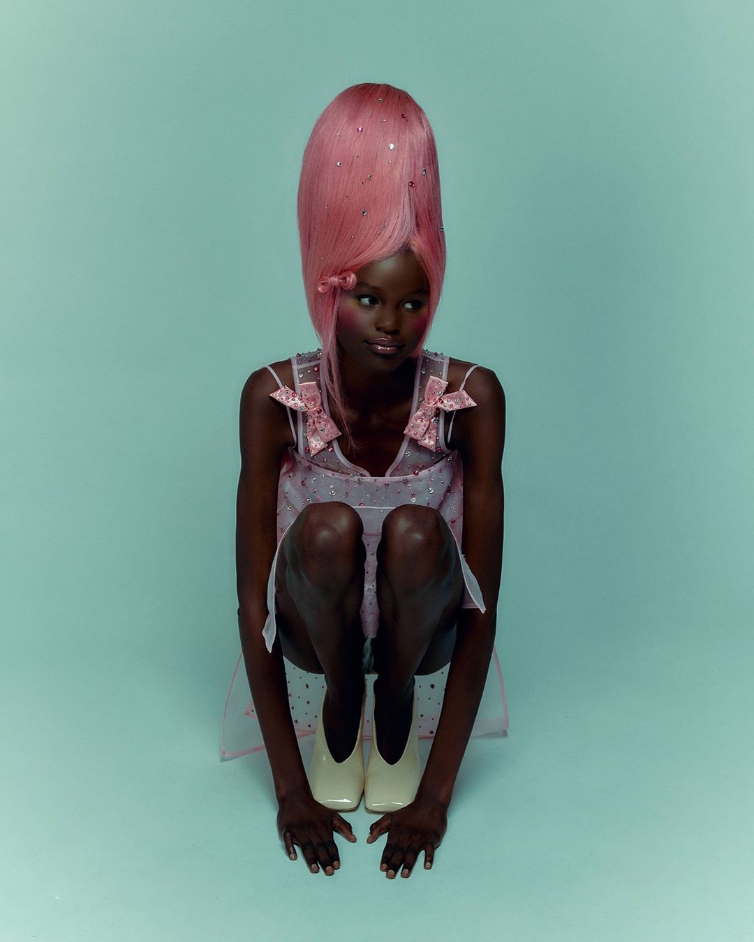 A dark-skinned woman with pink wig, wearing a pale pink dress and white shoes is crouching in a studio with light blue backdrop.
