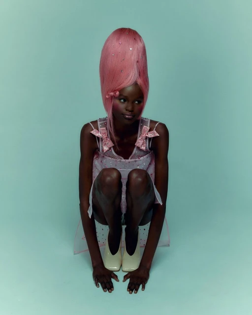 A dark-skinned woman with pink wig, wearing a pale pink dress and white shoes is crouching in a studio with light blue backdrop.