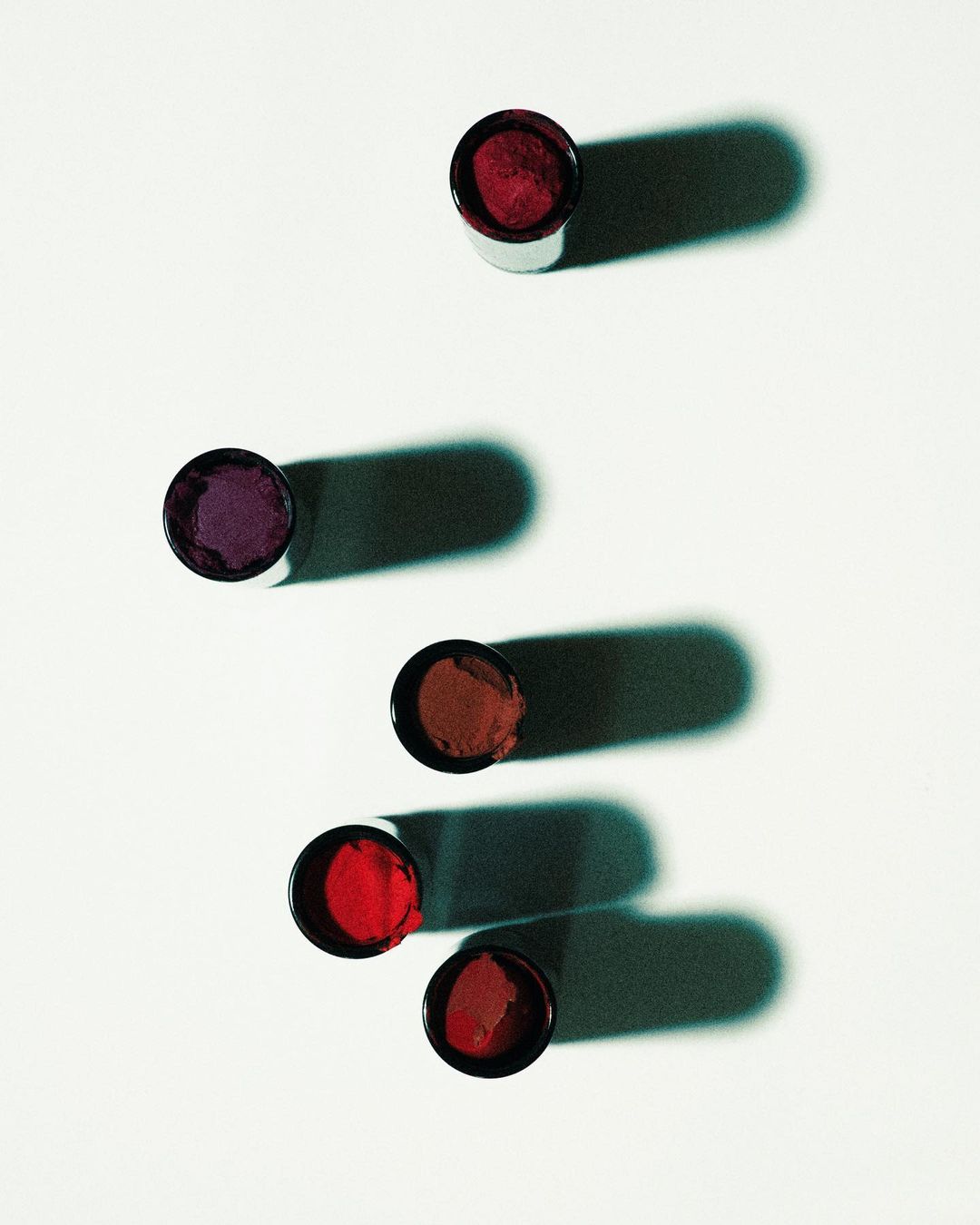 Several open lip gloss containers of various shades of red and purple are arranged on a white surface with shadows.