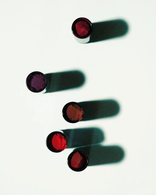 Several open lip gloss containers of various shades of red and purple are arranged on a white surface with shadows.