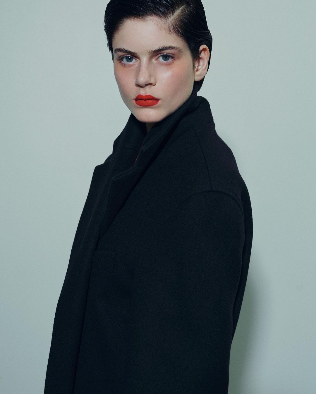 Portrait of a dark-haired model with bold red lips wearing a structured black coat against a pale grey background.