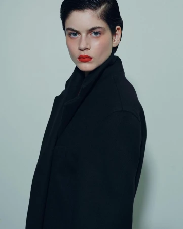 Portrait of a dark-haired model with bold red lips wearing a structured black coat against a pale grey background.
