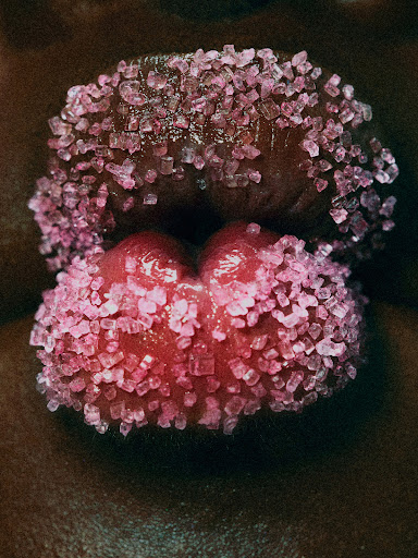 Macro close-up of a human mouth, lips covered in glittering pink sugar crystals. Dark skin tone frames the pink hues.