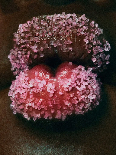 Macro close-up of a human mouth, lips covered in glittering pink sugar crystals. Dark skin tone frames the pink hues.