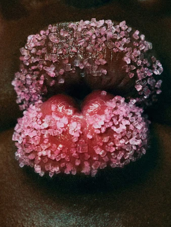 Macro close-up of a human mouth, lips covered in glittering pink sugar crystals. Dark skin tone frames the pink hues.