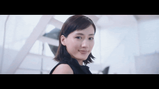 Smiling Asian woman in black top stands indoors with geometric white background, looking at camera in bright, modern setting.