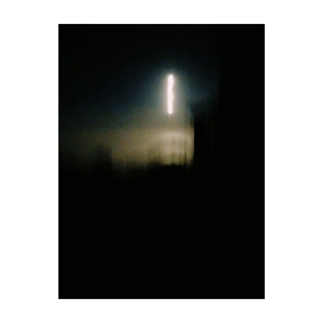 Abstract, blurred photograph of a vertical white light source on a dark background with a dark, indistinct structure in front.