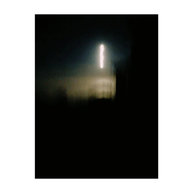 Abstract, blurred photograph of a vertical white light source on a dark background with a dark, indistinct structure in front.