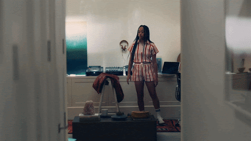 Young woman in a striped matching set stands in a living room, next to a coffee table and stereo equipment, viewed through a doorway.